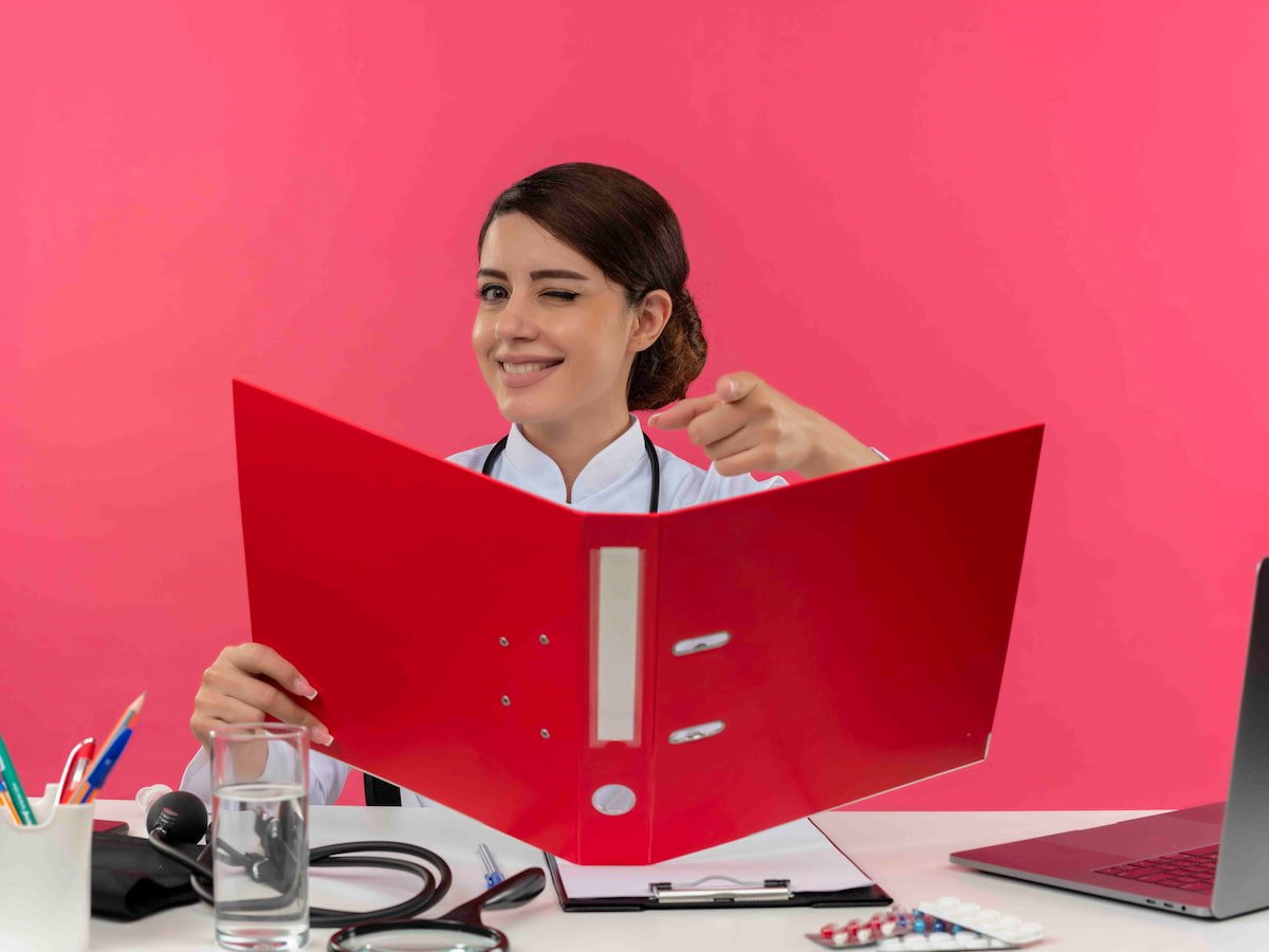 A female medical student smiling while holding a red folder and pointing toward the viewer, with a stethoscope on the desk and a bright pink background.
