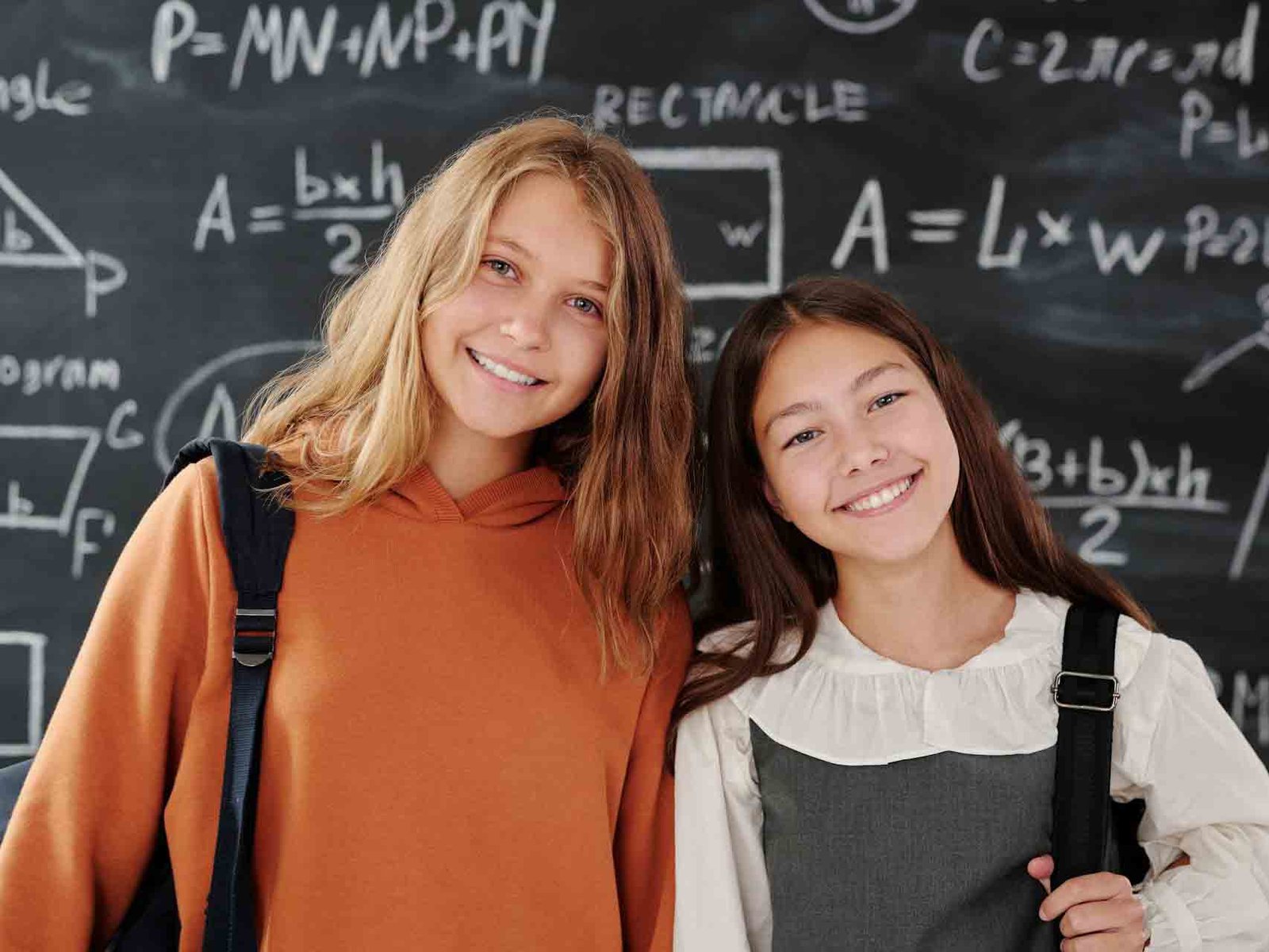 Two smiling schoolgirls standing together in front of a chalkboard with mathematical formulas, symbolizing the importance of girls’ education.