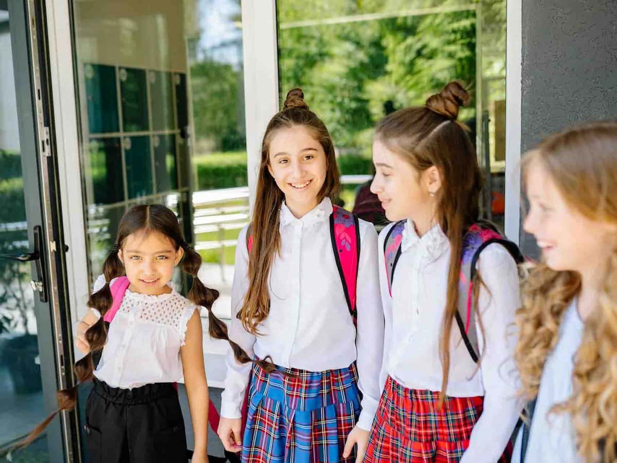 Four smiling schoolgirls standing outside a classroom, wearing backpacks and school uniforms, symbolizing the role of girls' education in shaping communities.